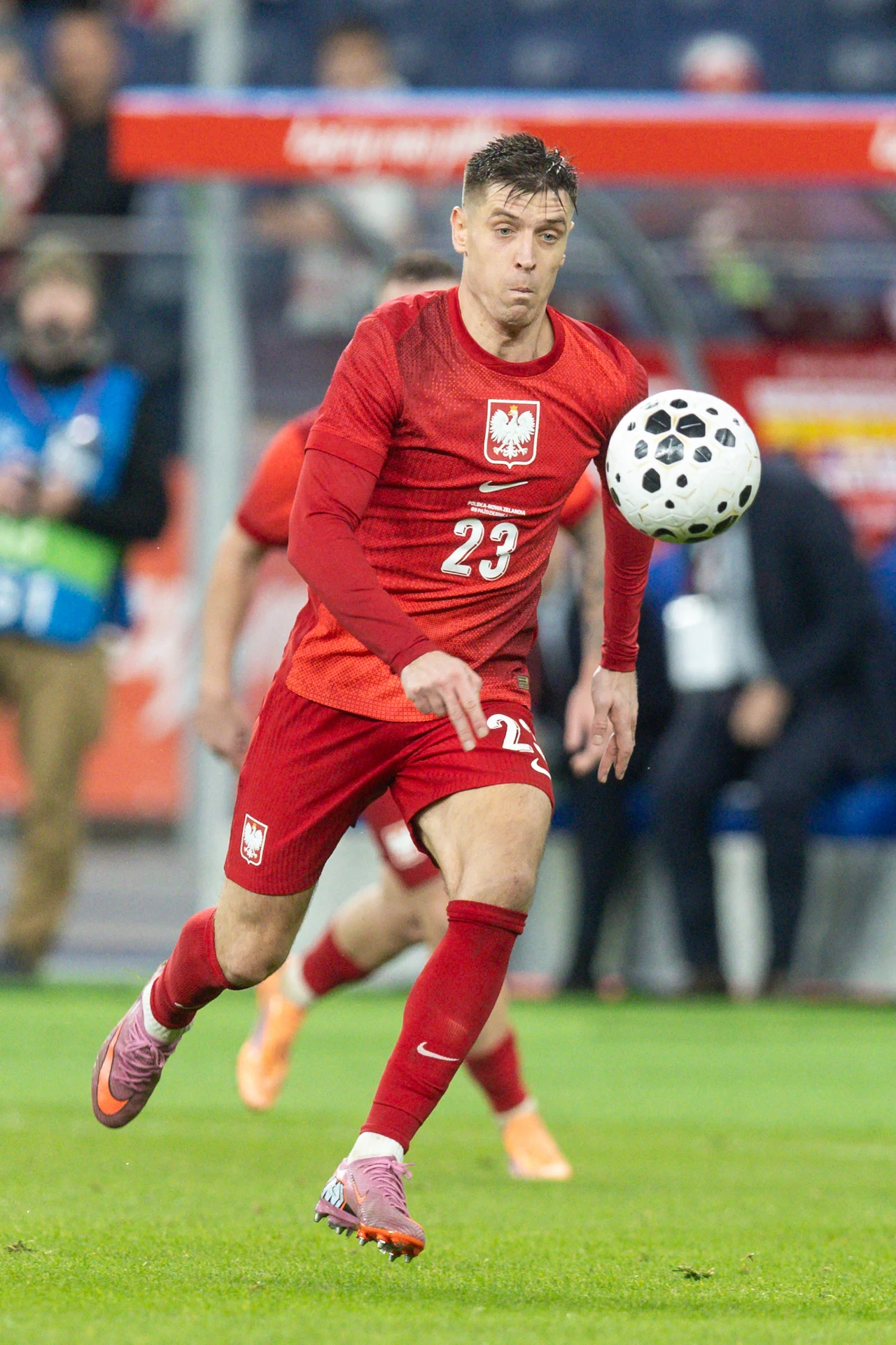 Polski piłkarz w czerwonym stroju z numerem 23 biegnie za piłką na murawie podczas meczu, w tle widoczni są inni zawodnicy i osoby obsługi technicznej.