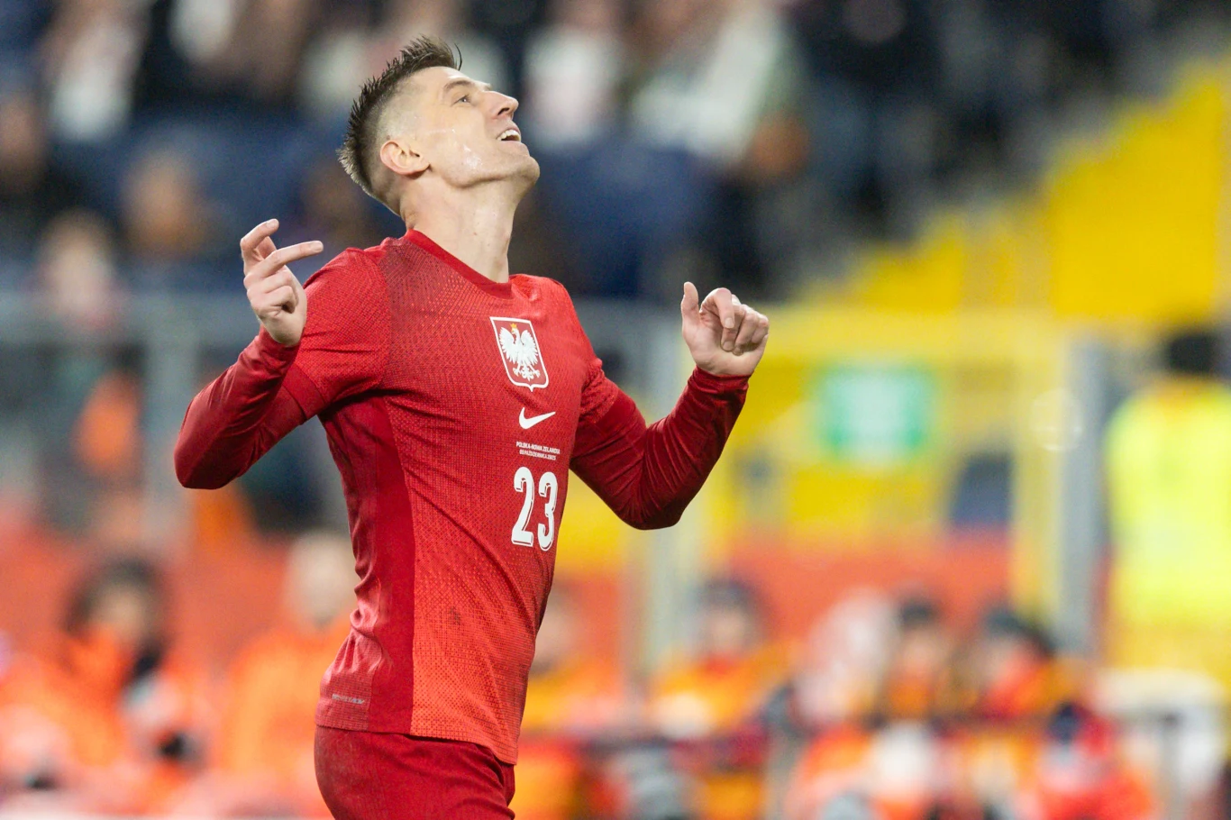 Krzysztof Piątek Piłkarz w czerwonym stroju reprezentacji Polski z numerem 23 unosi głowę i ręce do góry, wyrażając emocje podczas meczu, w tle rozmazani kibice i elementy stadionu.