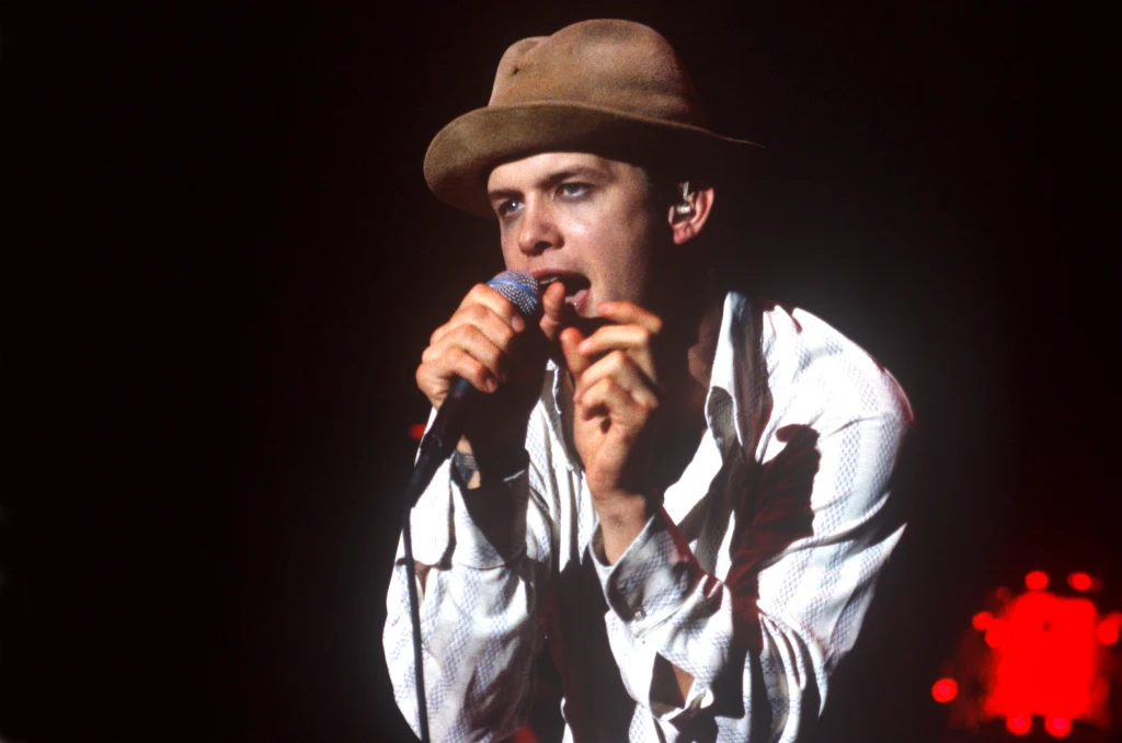 Mężczyzna w jasnej koszuli i brązowym kapeluszu śpiewający do mikrofonu na scenie, intensywne światło skierowane na postać, w tle rozmyte czerwone światła.