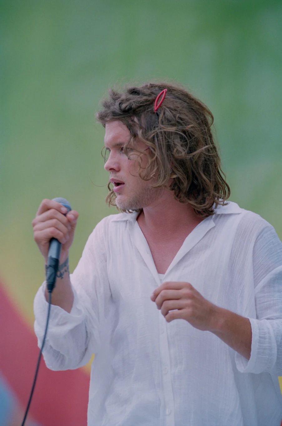 Mężczyzna z długimi, potarganymi włosami spiętymi spinką, ubrany w białą koszulę, trzyma mikrofon i śpiewa na tle rozmytego, zielonego tła.