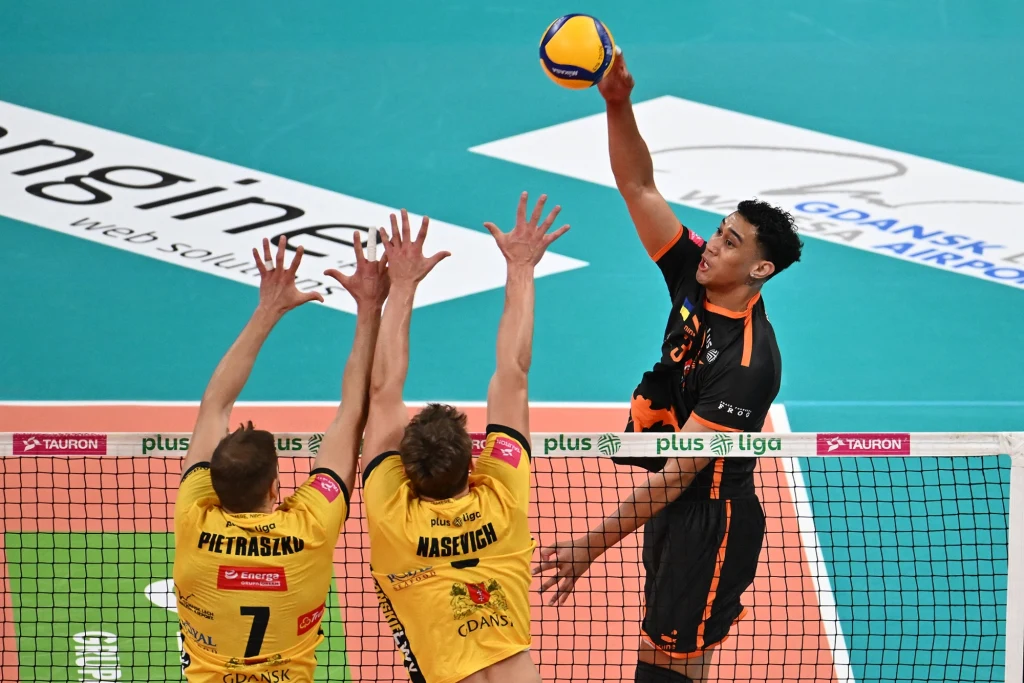 Lorenzo Pope w ataku w meczu Energa Trefl - Barkom Każany Atakujący w czarno-pomarańczowym stroju wybija piłkę siatkową ponad siatką, próbując przełamać blok dwóch zawodników w żółtych koszulkach z drużyny przeciwnej. W tle widoczna reklama i fragment boiska do siatkówki.