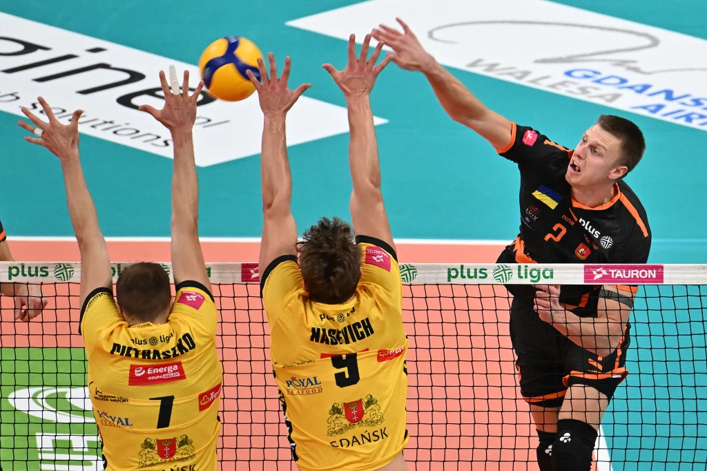 W ataku Ilja Kowaliow z Barkomu Każany Lwów Zawodnik w czarnym stroju atakuje piłkę siatkową ponad siatką, dwóch zawodników w żółtych koszulkach próbuje zablokować atak, widoczne logo Plus Liga na siatce, akcja rozgrywa się na hali sportowej.