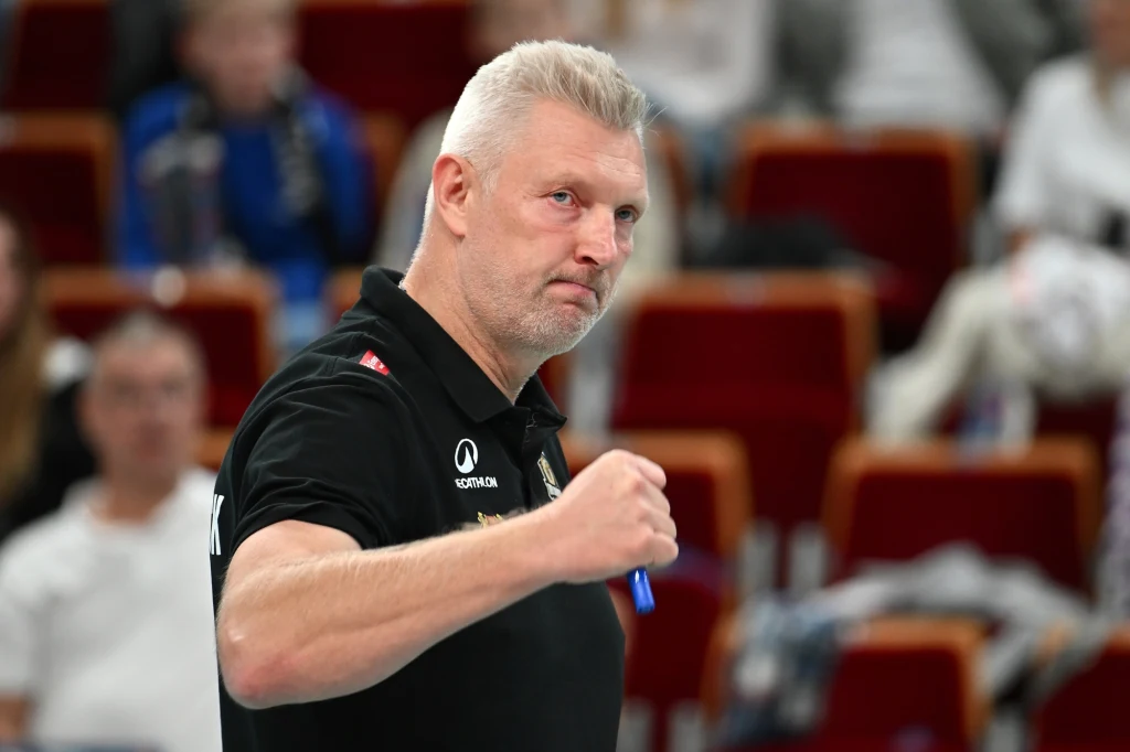 Mariusz Sordyl, trener Energa Trefla Gdańsk Mężczyzna z krótkimi siwymi włosami w czarnej koszulce z uniesioną ręką w geście triumfu, siedzący na tle rozmazanych czerwonych foteli w hali sportowej.