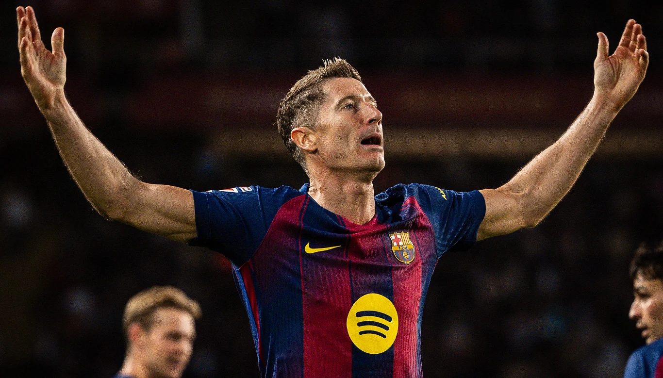 Robert Lewandowski Piłkarz w stroju FC Barcelony z uniesionymi ramionami, wyrażający emocje podczas meczu, w tle rozmyte sylwetki innych graczy i trybun.