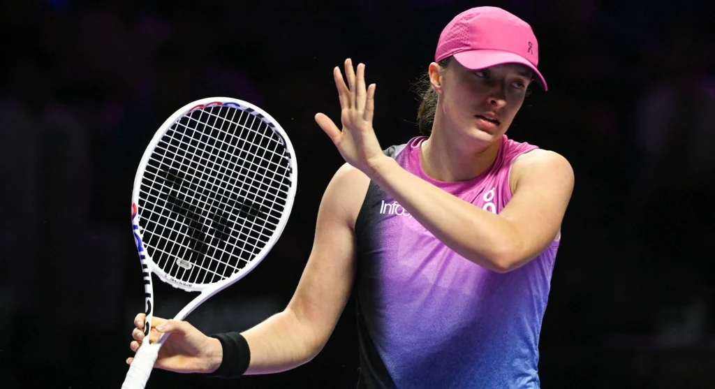 Tenisistka w różowej czapce i sportowej koszulce trzyma rakietę tenisową, gestykulując dłonią podczas intensywnej gry na korcie, tło jest ciemne, skupienie na sportowcu.