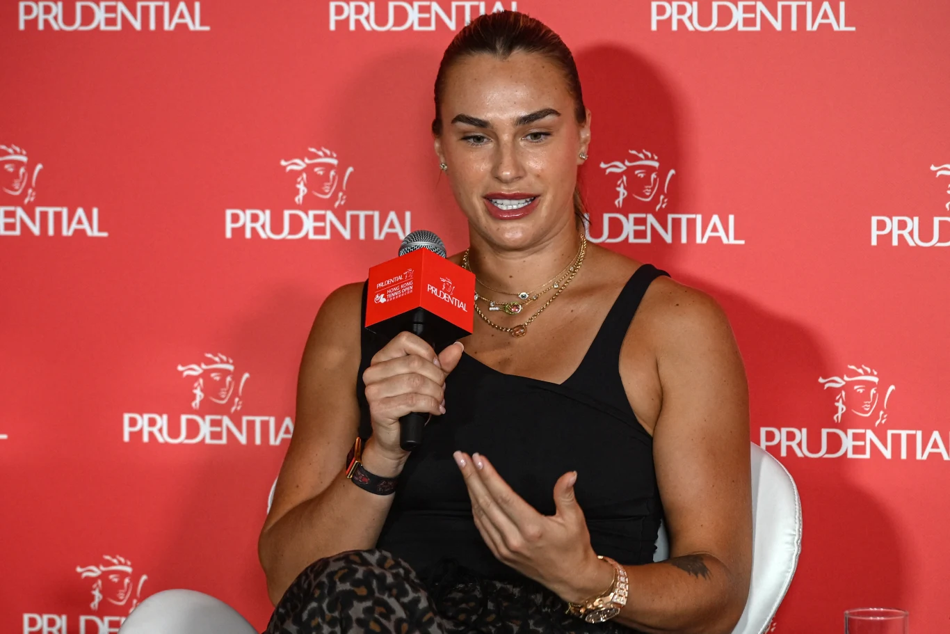 Aryna Sabalenka Kobieta wypowiadająca się podczas konferencji prasowej, trzyma mikrofon z logo Prudential, w tle czerwony baner z logotypem tej firmy, ubrana w czarną koszulkę oraz kilka naszyjników i bransoletek.