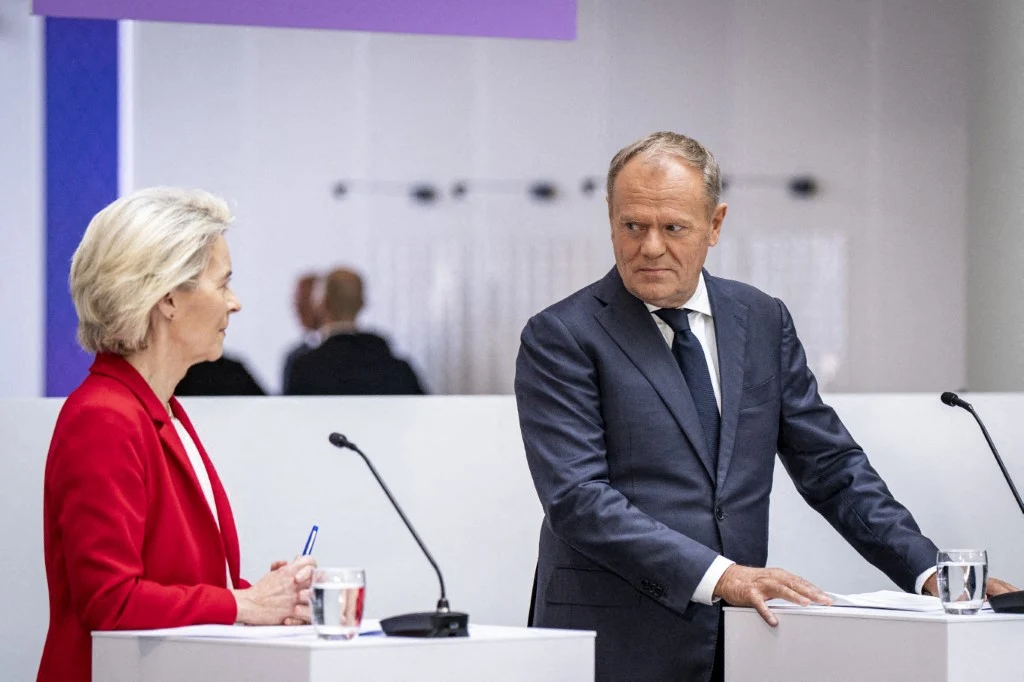 Co dalej z ETS2? Decyzja już została podjęta Kobieta w czerwonym żakiecie oraz mężczyzna w granatowym garniturze stoją przy białych pulpitach, prowadząc rozmowę podczas wystąpienia lub konferencji prasowej. Przed nimi znajdują się mikrofony i szklanki z wodą, a w tle widoczne są rozmyte postacie ...