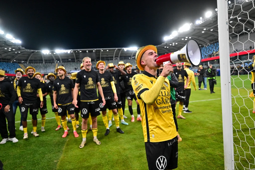 Piłkarz w żółto-czarnym stroju przemawia do megafonu stojąc przy bramce, w tle grupa uśmiechniętych zawodników tej samej drużyny świętuje na stadionie przy sztucznym oświetleniu