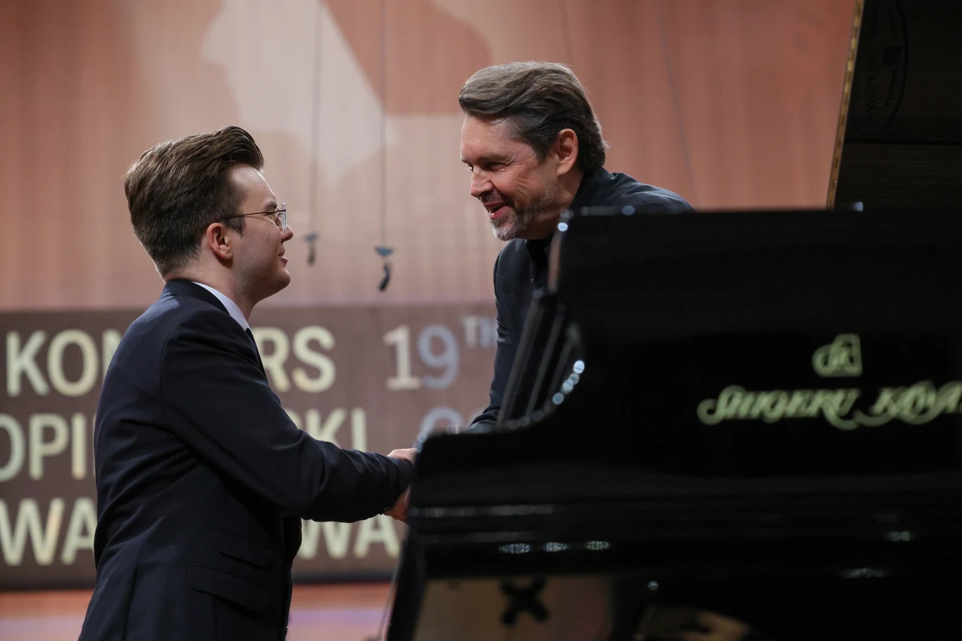 Piotr Alexewicz (z lewej) oraz dyrygent Konkursu Chopinowskiego Andrzej Boreyko Dwóch mężczyzn w eleganckich strojach ściska sobie dłonie na scenie koncertowej obok fortepianu, w tle widoczny fragment napisu sugerujący prestiżowe wydarzenie muzyczne.
