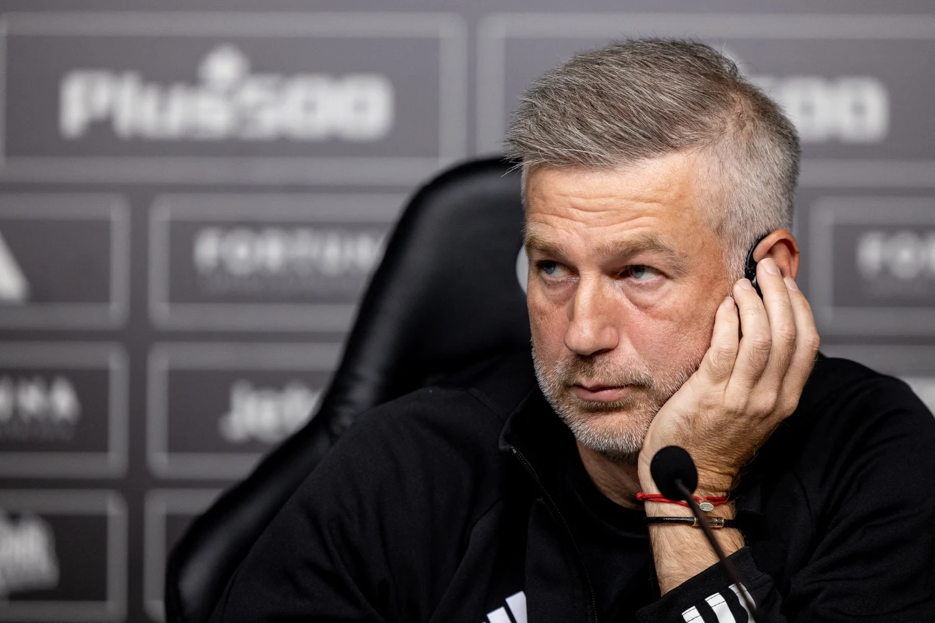 Mężczyzna z siwiejącymi włosami siedzi przy mikrofonie, opiera głowę o dłoń i słucha uważnie podczas konferencji prasowej. W tle widoczne zamazane logo sponsorów oraz czarno-białe tło.