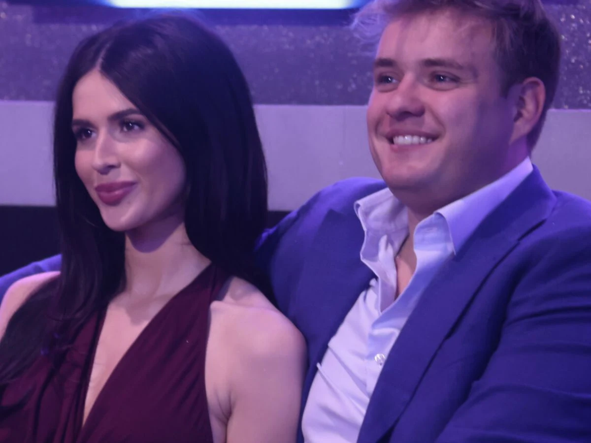 Roksana Węgiel i Kevin Mglej Para siedząca obok siebie, uśmiechnięci, ubrani elegancko – kobieta w bordowej sukience, mężczyzna w jasnej koszuli i niebieskiej marynarce, w tle dekoracja w odcieniach fioletu.