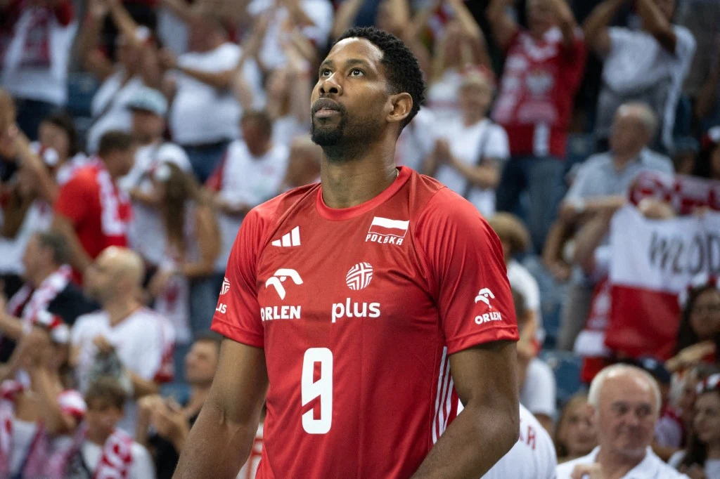 Wilfredo Leon Siatkarz w czerwonym stroju z numerem 9 stoi na hali sportowej w otoczeniu kibiców w białych i czerwonych barwach, w tle widoczne transparenty i tłum ludzi.