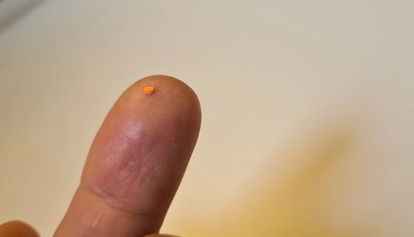 Takie maleństwo, a tyle zmienia. Tak wygląda implant Prima Mała pomarańczowa tabletka lub drażetka umieszczona na opuszku palca wskazującego na tle jednolitej, jasnej powierzchni.