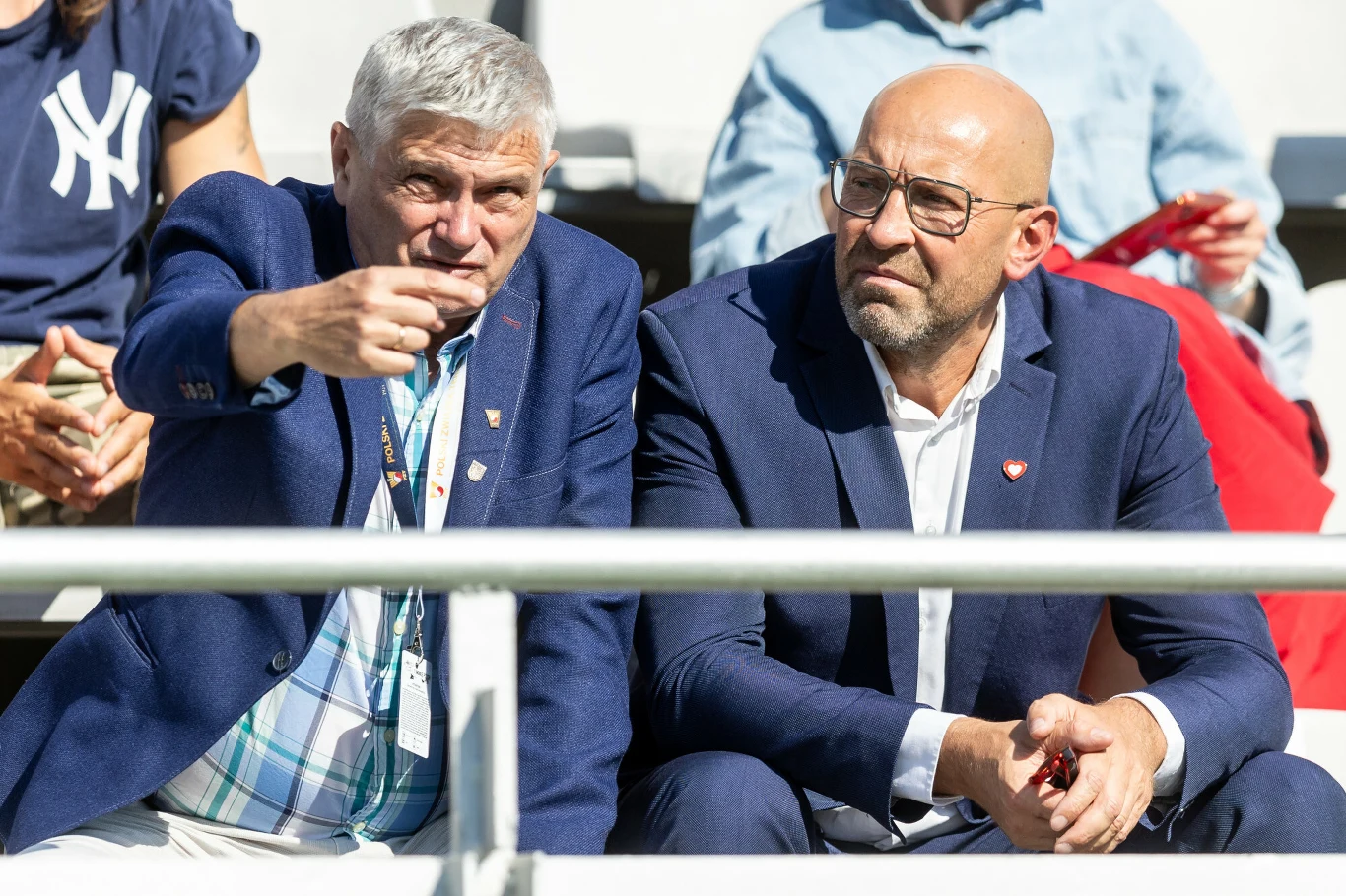 Dwóch mężczyzn w garniturach siedzi na trybunach stadionu, jeden z nich gestykuluje ręką wskazując coś przed sobą, obaj są skupieni i zaangażowani w obserwowanie wydarzenia.