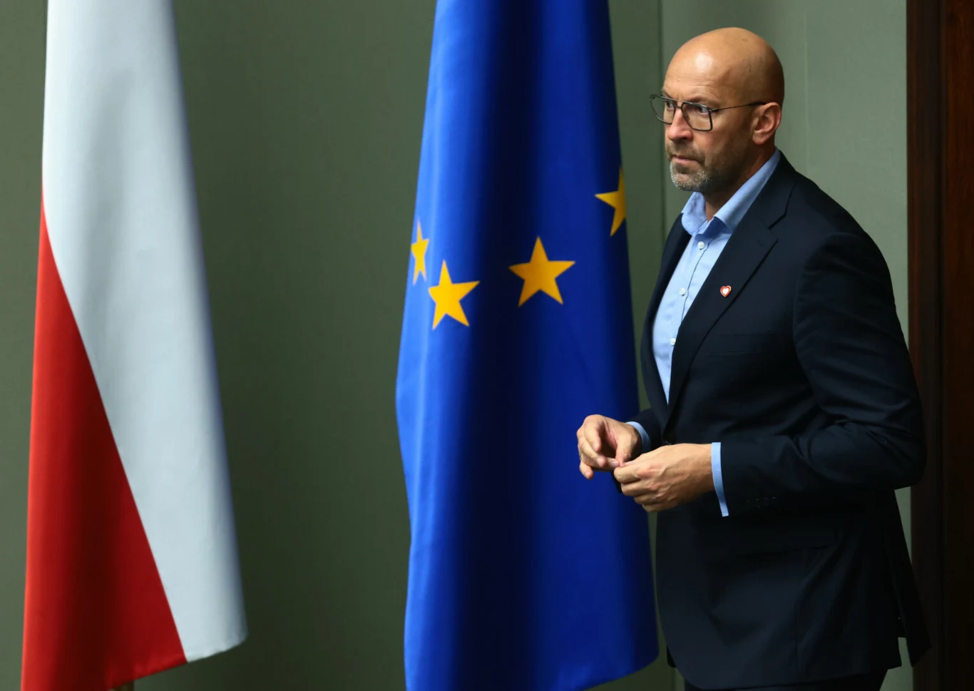 Jakub Rutnicki, 10.09.2025 r. Mężczyzna w garniturze i okularach stoi obok flagi Polski oraz flagi Unii Europejskiej, ma poważny wyraz twarzy i znajduje się w zamkniętym pomieszczeniu.