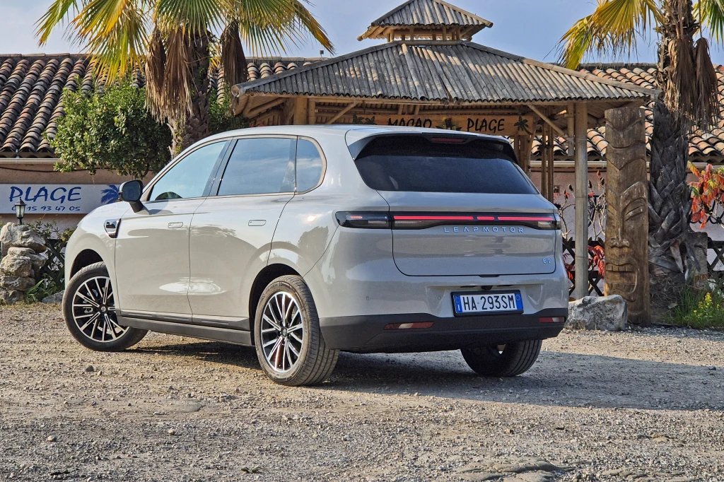 Z tej perspektywy widać podobieństwa B10 do większego modelu marki, czyli C10 Srebrny samochód elektryczny zaparkowany na żwirowym parkingu przed drewnianą altaną ozdobioną palmami.