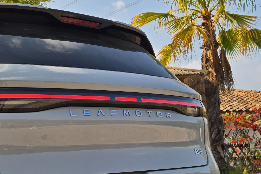 Leapmotor B10 Tył srebrnego samochodu elektrycznego marki Leapmotor z czerwonym pasem świetlnym oraz literami logo na klapie bagażnika, w tle palma i fragment dachu wraz z niebieskim niebem.