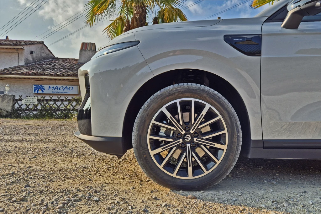 Leapmotor B10 Przednie koło samochodu na żwirowej drodze, widoczna elegancka felga oraz fragment maski i reflektora auta, w tle dom z palmą i banerem z napisem Macao.