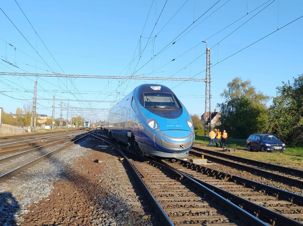 Nowoczesny niebieski pociąg typu Pendolino stojący na torach kolejowych, grupa pracowników w odblaskowych kamizelkach obserwuje sytuację z boku, na poboczu widoczny zaparkowany samochód oraz sieć trakcyjna nad torami.