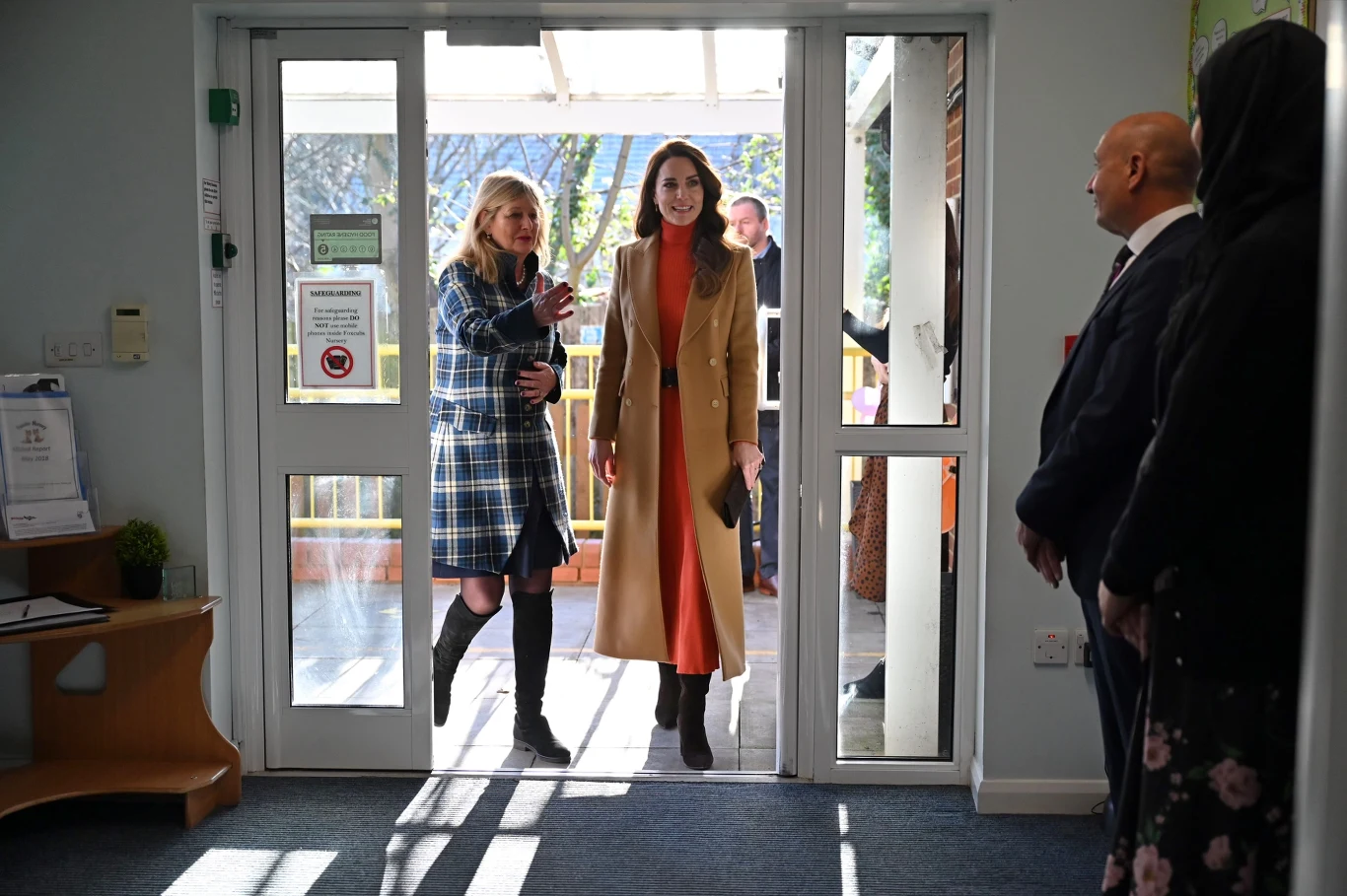 Księżna Kate zachwyciłą tym zestawem w 2023 roku i jest on wciąż w obowiązujących na ten rok trendach Dwie kobiety wchodzą do budynku przez przeszklone drzwi, jedna ubrana w płaszcz w kratę z czarną torbą, druga w długi camelowy płaszcz z pomarańczową sukienką, w środku przy wejściu stoi dwóch mężczyzn i kobieta w ciemnym płaszczu.