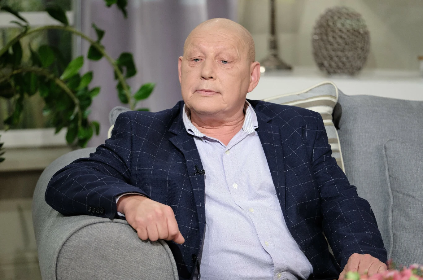 Krzysztof Jackowski zabrał głos w sprawie pogody na Wszystkich Świętych Łysy mężczyzna w eleganckiej marynarce siedzi na jasnej sofie, w tle jasne wnętrze z rośliną doniczkową i dekoracyjnymi poduszkami.