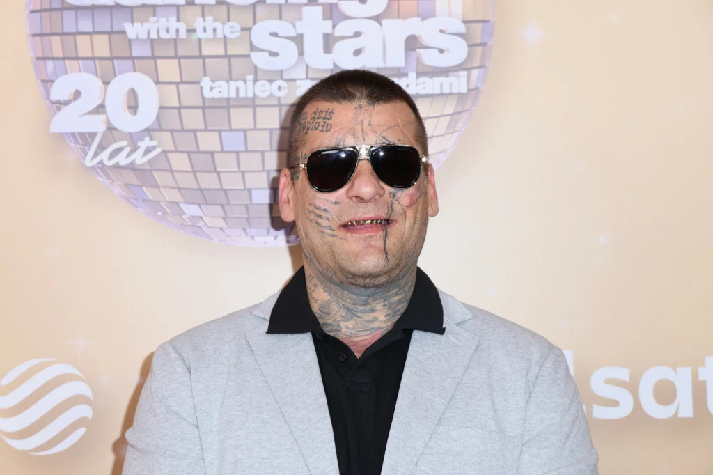 Paweł Mikołajuw "Popek" Mężczyzna w jasnej marynarce i czarnej koszuli, z widocznymi tatuażami na twarzy i szyi oraz w okularach przeciwsłonecznych, stojący na tle ścianki promocyjnej programu tanecznego.