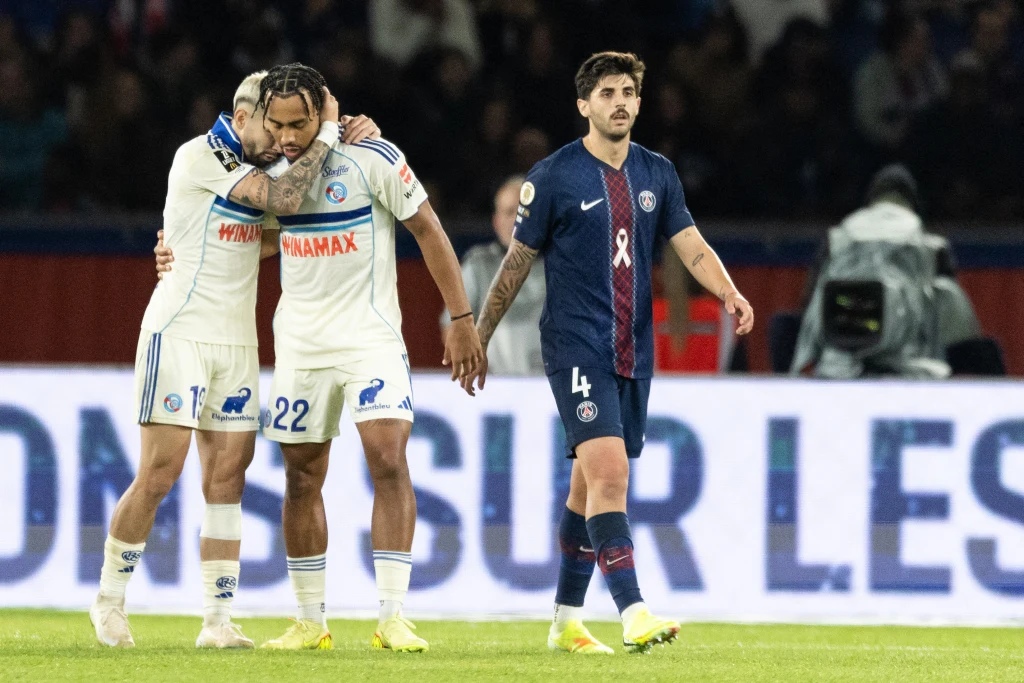 Piłkarze RC Strasbourg w zremisowanym 3:3 meczu Ligue 1 z PSG Piłkarze w strojach klubowych znajdują się na boisku, dwóch zawodników drużyny w białych strojach wspiera się wzajemnie, trzeci piłkarz w granatowym stroju przechodzi obok nich, w tle rozmazana publiczność.