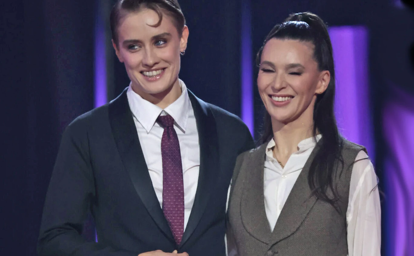 Katarzyna Zillmann i Janja Lesar w "Tańcu z gwiazdami" Dwie kobiety stoją blisko siebie na scenie, obie są ubrane elegancko, jedna w garnitur z krawatem, druga w biały koszuli i kamizelce, na ich twarzach widoczne są uśmiechy, a tło oświetlone jest fioletowym światłem.