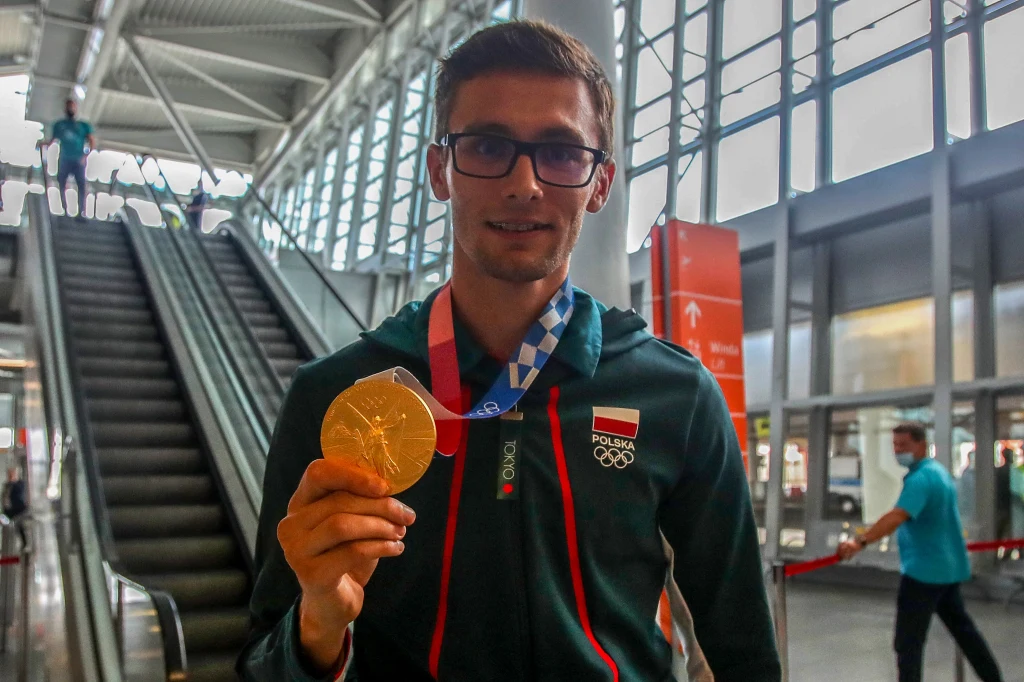 Mężczyzna w stroju reprezentacji Polski stojący na tle lotniska trzyma w ręku złoty medal olimpijski, uśmiechając się do obiektywu.