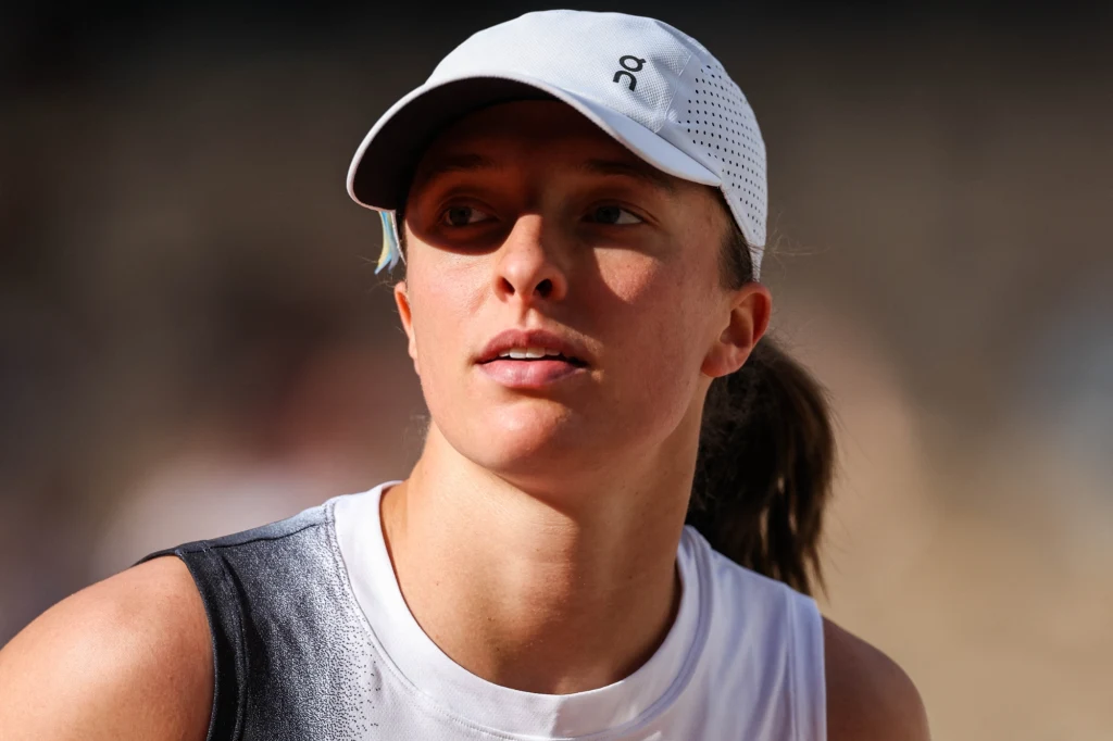 Iga Świątek Młoda kobieta w sportowej koszulce i jasnej czapce z daszkiem, skierowana twarzą w bok, w naturalnym świetle na tle rozmytego tła.