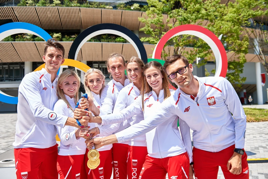 Grupa polskich sportowców ubranych w białe dresy z czerwonymi akcentami prezentuje zdobyte złote medale na tle kolorowych kół olimpijskich i nowoczesnego obiektu sportowego.
