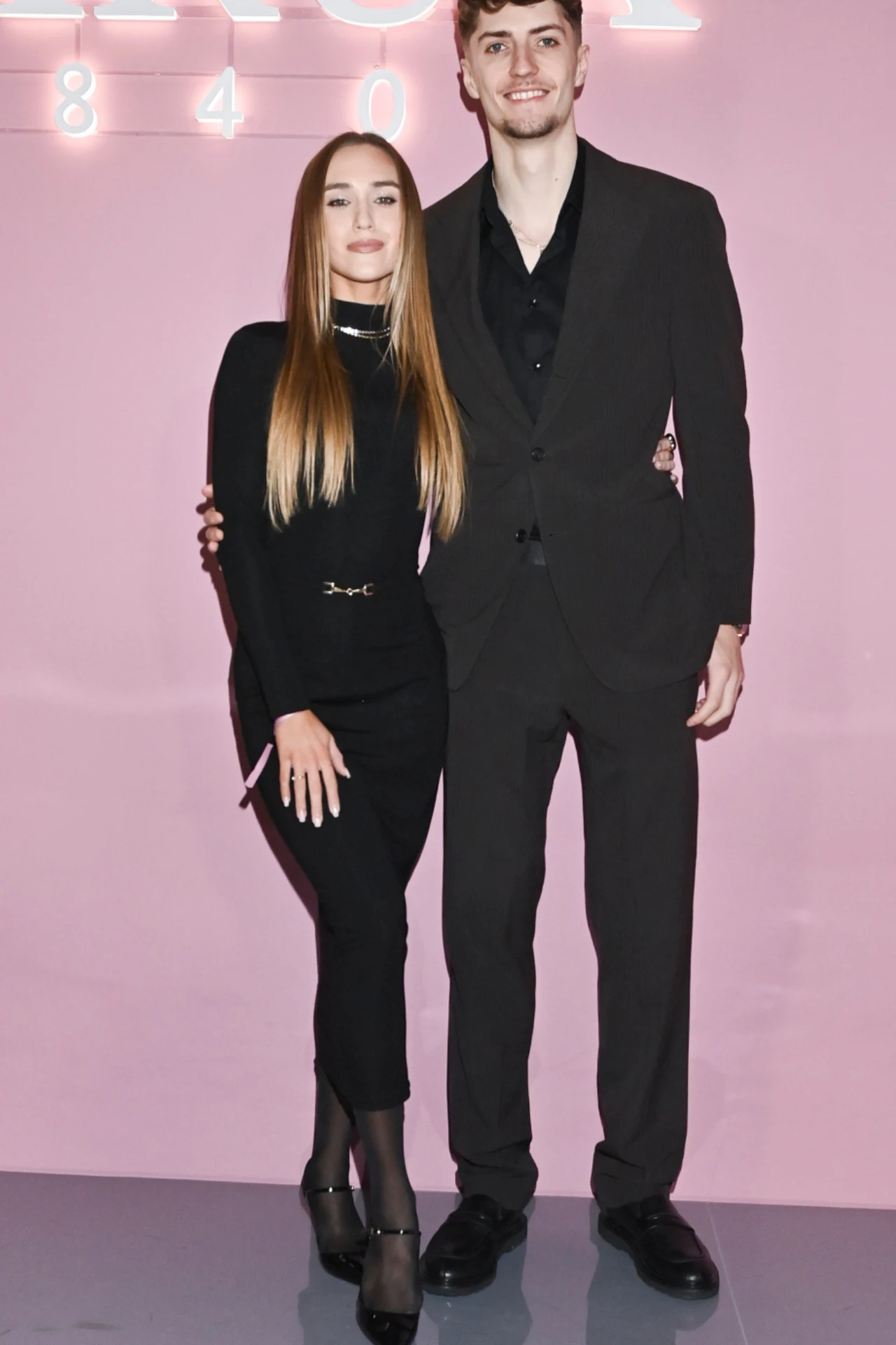 Daria Syta i Igor Grobelny na wydarzeniu branżowym Młoda kobieta i mężczyzna w eleganckich, ciemnych stylizacjach stoją razem na tle jasnoróżowej ściany z podświetlonym napisem, trzymając się za ramiona i uśmiechając się delikatnie.