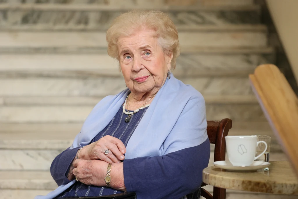 Starsza kobieta o blond włosach i pogodnym wyrazie twarzy siedzi na krześle przed marmurowymi schodami, ubrana w niebieską suknię i jasny szal, na stoliku obok stoi filiżanka z napojem.