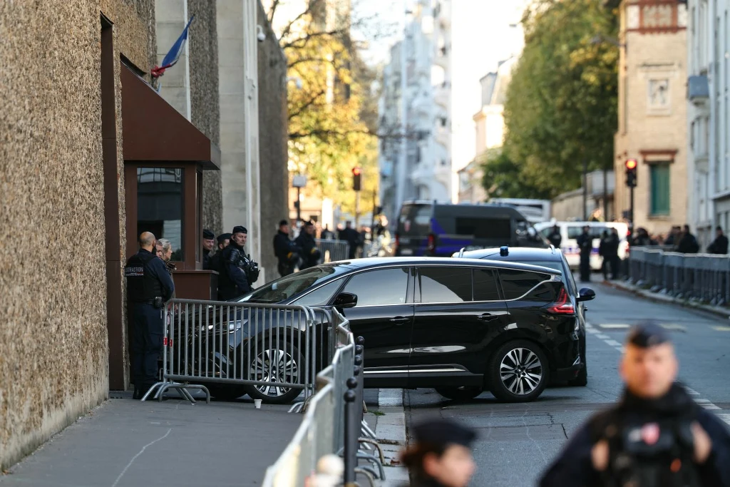 Czarny samochód osobowy zatrzymany przy barierekach ochronnych, wokół kilku policjantów zabezpieczających wjazd do budynku, ulica ogrodzona metalowymi zaporami, w tle widocznych więcej funkcjonariuszy.