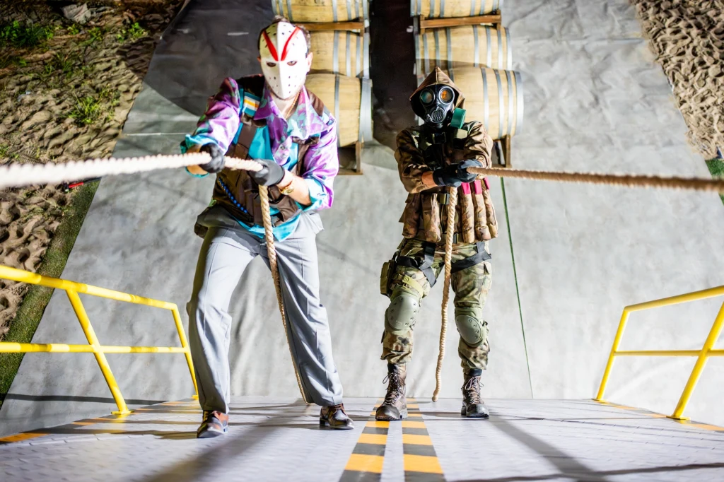 Dwie osoby w maskach przeciągają linę na stromym podjeździe otoczonym żółtymi barierkami, jedna ubrana w kolorową kurtkę z maską, druga w militarnej odzieży z maską gazową, w tle widoczne worki na piasku.