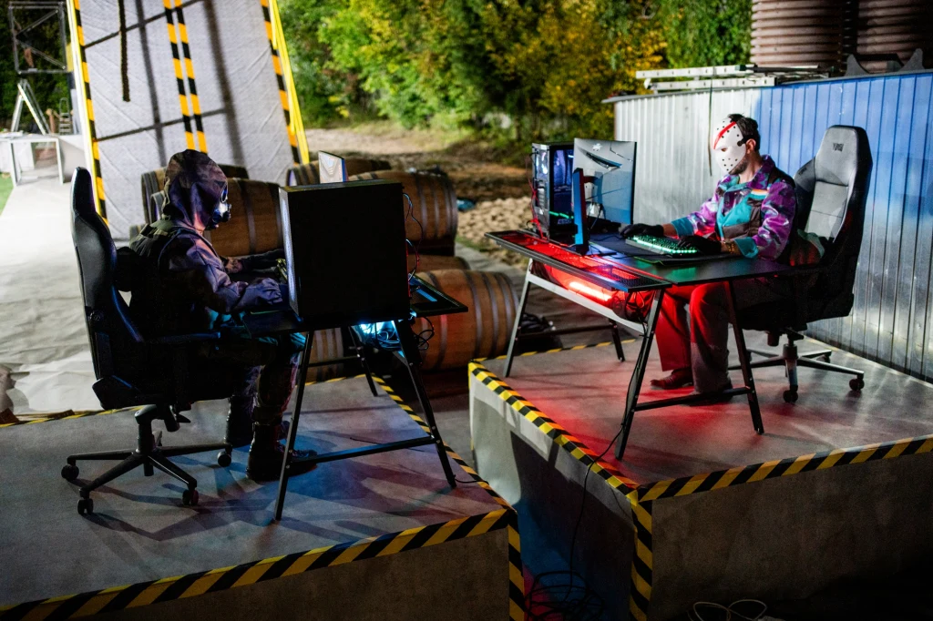 Dwóch graczy w maskach i kolorowych strojach siedzi przy komputerach na betonowych platformach otoczonych żółto-czarnymi taśmami ostrzegawczymi, obok widać beczki i metalowy płot, w tle zieleń drzew.