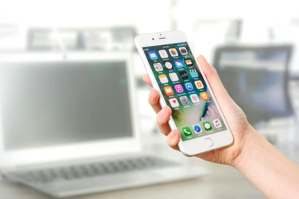 Apki dietetyczne na iOS Smartfon trzymany w dłoni na tle rozmytego ekranu laptopa, wyświetlający ekran główny z aplikacjami.