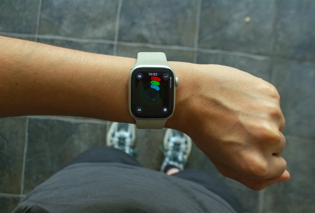 Najlepsze apki dietetyczne w Polsce Smartwatch na lewym nadgarstku, ekran urządzenia wyświetla kolorową grafikę przedstawiającą aktywność fizyczną, tło stanowi szara posadzka, widoczne są fragmenty nóg osoby w sportowym stroju i butach.