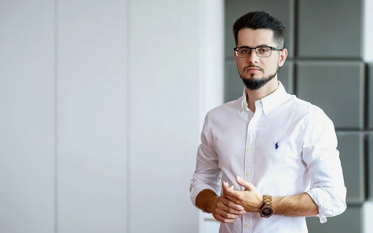 Adrian Pakulski, współwłaściciel spółki Enduro7, zarządzającej marką Motogar Polska Mężczyzna z brodą i okularami w białej koszuli z długim rękawem stoi w jasnym, nowoczesnym wnętrzu, patrząc w stronę obiektywu.