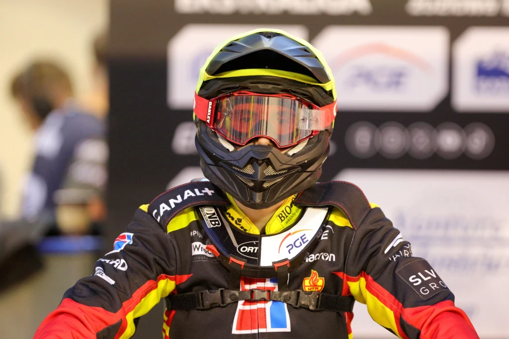 William Drejer w barwach Sparty Wrocław. Zawodnik ubrany w kolorowy strój żużlowy z hełmem i goglami, na tle rozmazanych banerów sponsorów. W centrum kadru skoncentrowany jest na sportowcu, wszystkie elementy stroju wskazują na udział w zawodach motorowych.