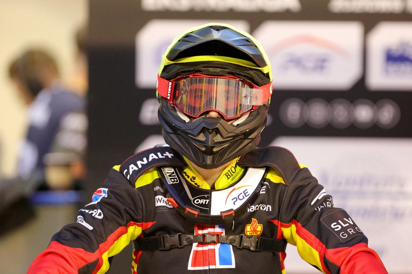 William Drejer w barwach Sparty Wrocław. Zawodnik ubrany w kolorowy strój żużlowy z hełmem i goglami, na tle rozmazanych banerów sponsorów. W centrum kadru skoncentrowany jest na sportowcu, wszystkie elementy stroju wskazują na udział w zawodach motorowych.