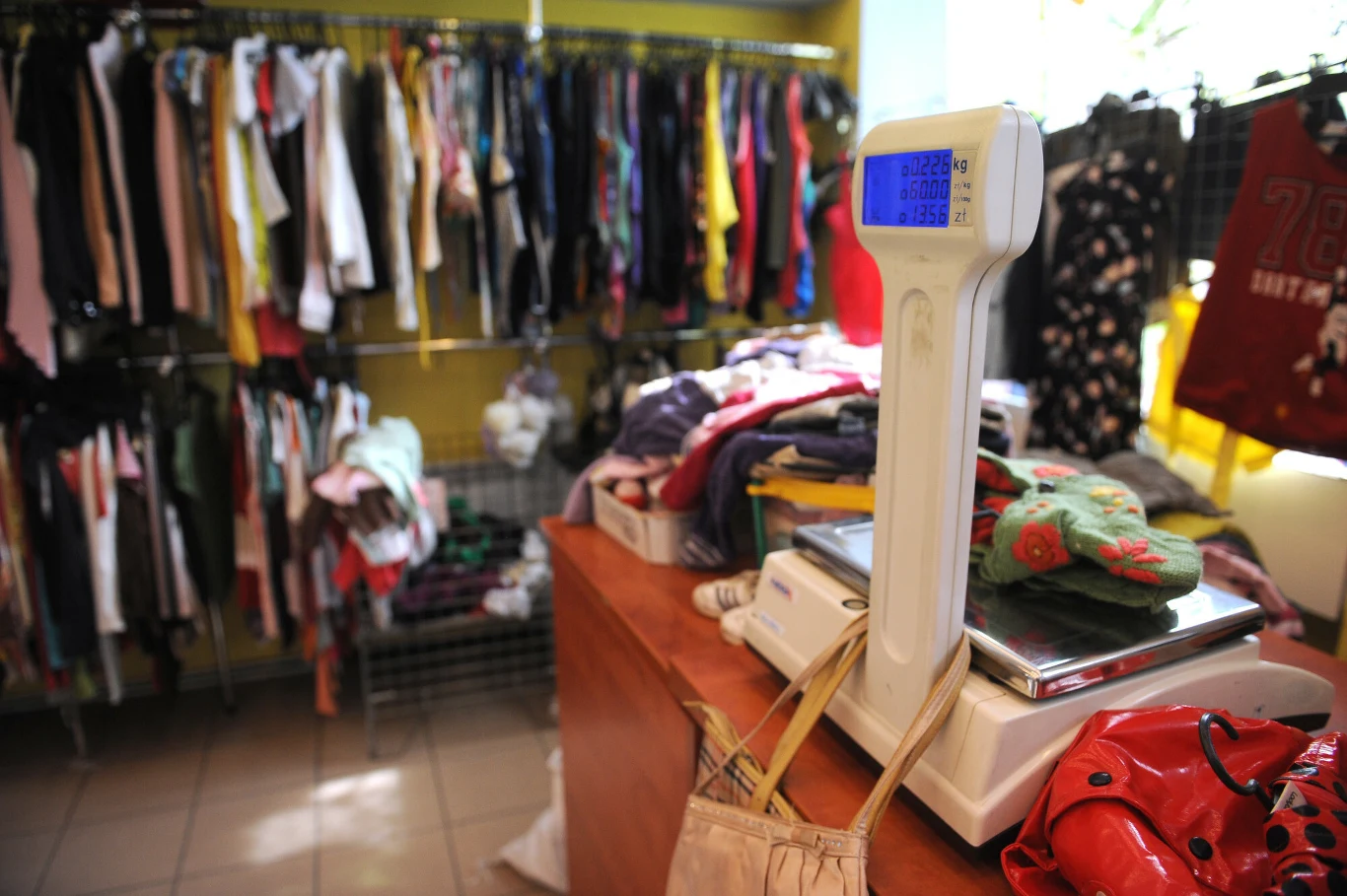 Stos ubrań na wadze elektronicznej ustawionej na ladzie, w tle widoczne wieszaki z różnorodną odzieżą używaną zawieszoną na metalowych kratkach oraz jasne wnętrze sklepu
