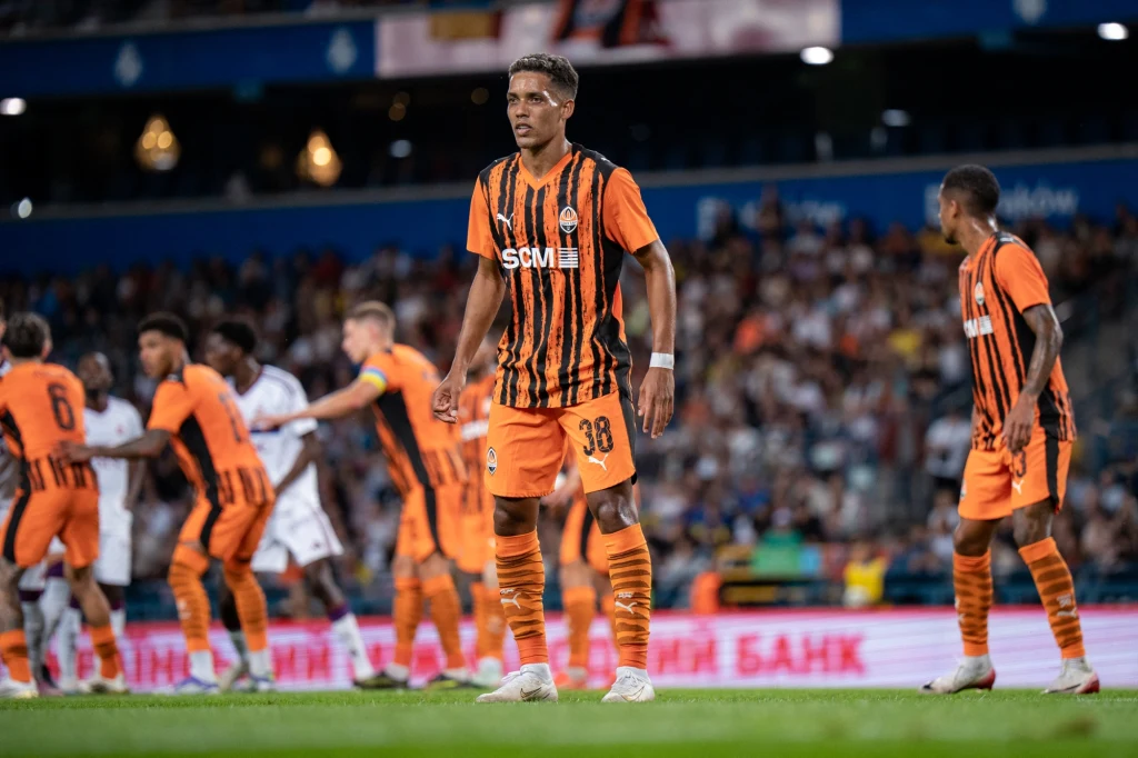 Piłkarze w pomarańczowo-czarnych strojach na boisku podczas meczu, w tle rozmyte sylwetki zawodników rywali oraz trybuny pełne kibiców.