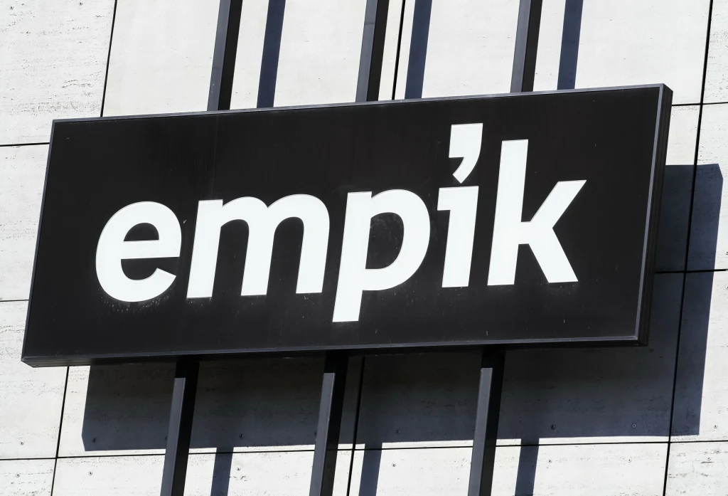 Czarno-biała tablica z logo sklepu Empik zamontowana na elewacji budynku, tło z jasnego betonu z pionowymi ciemnymi liniami.