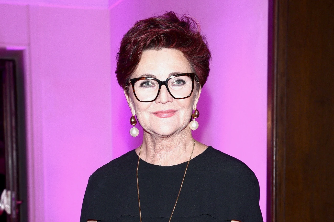 Jolanta Kwaśniewska słynie z doskonałego poczucia stylu. W stylizacjach Łukasza Jemioła prezentuje się fenomenalnie Kobieta w średnim wieku z krótkimi, ciemnoczerwonymi włosami, nosząca wyraziste okulary w czarnej oprawie, złote kolczyki z perłami oraz czarną bluzkę, stoi na tle oświetlonym fioletowym światłem.