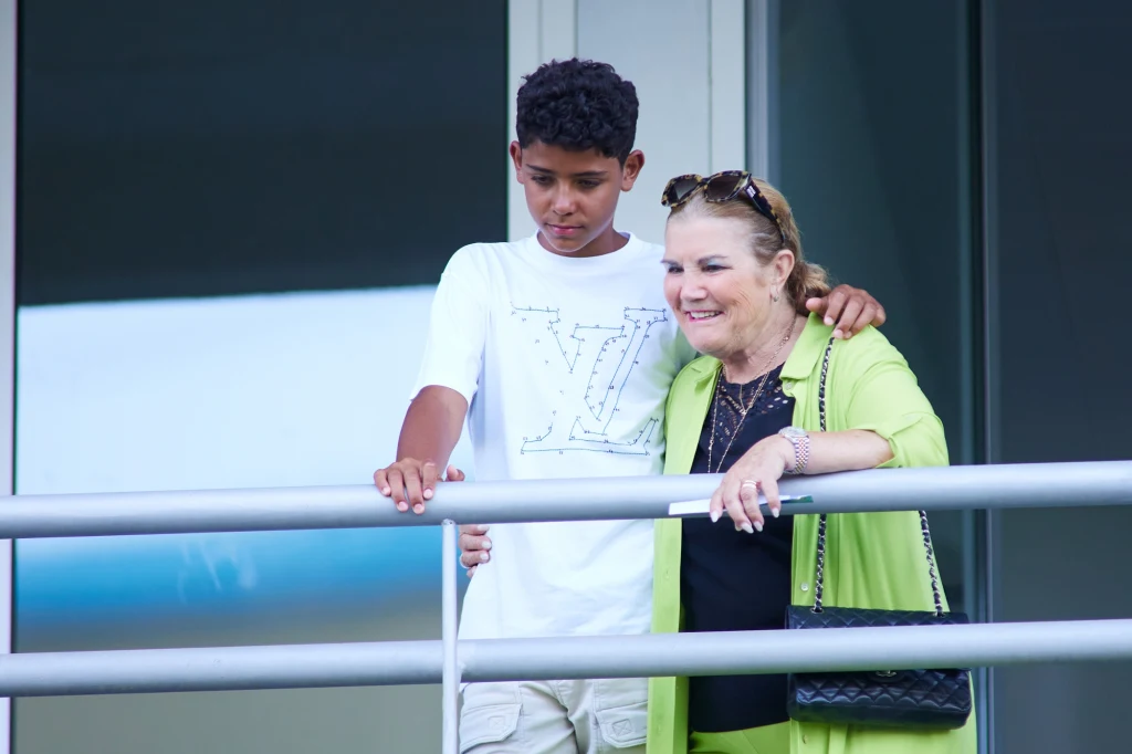 Matka i najstarszy syn Cristiano Ronaldo - Maria Dolores dos Santos Viveiros da Aveiro i Cristiano Junior Chłopiec i starsza kobieta stoją przy balustradzie balkonowej, uśmiechnięci, ubrani w jasne ubrania; kobieta w zielonym płaszczu, chłopiec ma biały t-shirt z dużym logotypem.