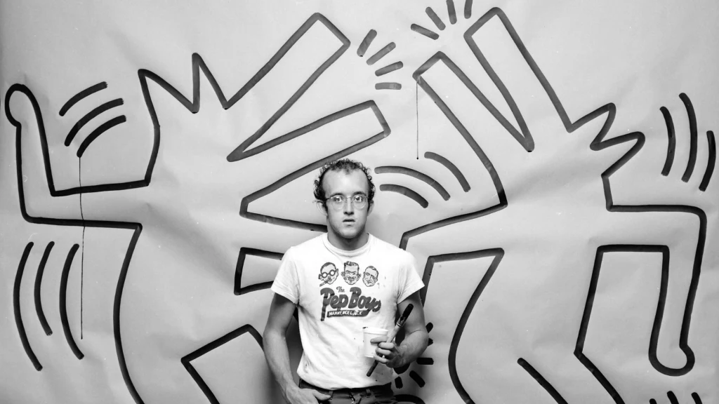 Keith Haring przed jedną ze swoich prac w 1983 roku Młody mężczyzna w okularach i koszulce z nadrukiem stoi na tle dużej ściany pokrytej charakterystycznymi czarnymi rysunkami abstrakcyjnych, tańczących postaci.