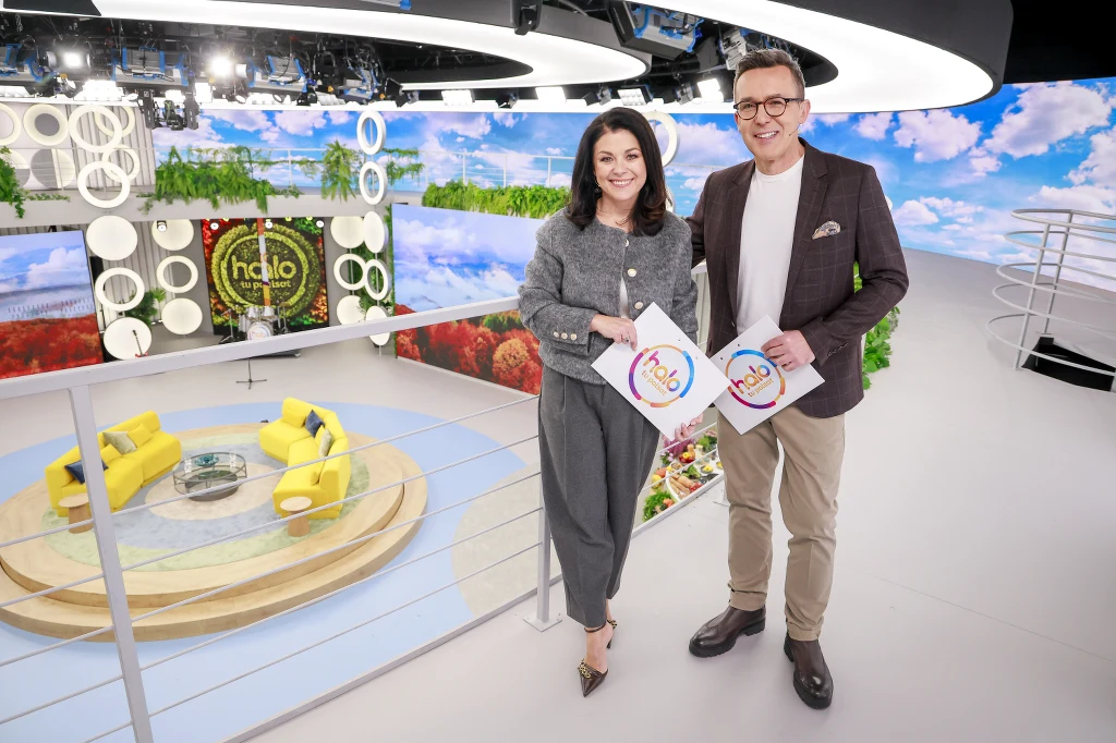 Katarzyna Cichopek i Maciej Kurzajewski na planie halo tu polsat Dwóch prezenterów uśmiechniętych, trzymających teczki z logo programu, stojących w nowoczesnym, kolorowym studiu telewizyjnym z żółtymi fotelami ustawionymi wokół szklanego stolika i dużym ekranem w tle pokazującym krajobraz z niebem i zielenią.