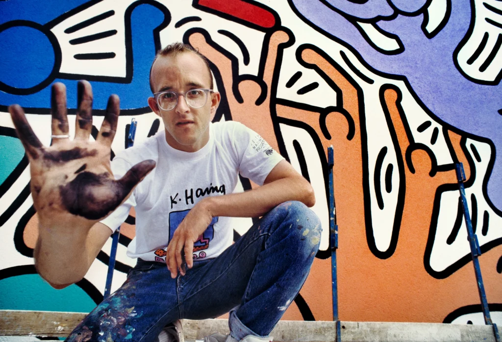 Keith Hering przed muralem "Tuttomondo" na ścianie kościoła Sant'Antonio w Pizie, 1989 r. Młody mężczyzna w okularach, ubrany w białą koszulkę i poplamione farbą jeansy, siedzi przed kolorowym muralem. Na tle graffiti unosi otwartą, pobrudzoną czarną farbą dłoń w kierunku obiektywu.