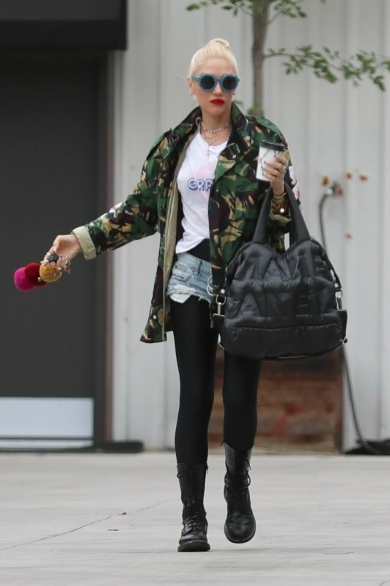 Kobieta w okularach przeciwsłonecznych i modnym stroju składającym się z militarnej kurtki, białej koszulki, szortów z dżinsu i czarnych legginsów, trzymająca dużą czarną torbę oraz napój na wynos. Jej stylizację uzupełniają czerwone usta i wysokie cza...